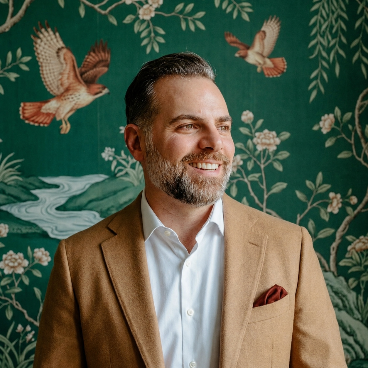 Tom Pepe: smiling headshot in a camel sport coat with a burnt umber pocket square, over a white shirt, standing before a deep viridian wallpaper patterned with red-tailed hawks, water, and florals